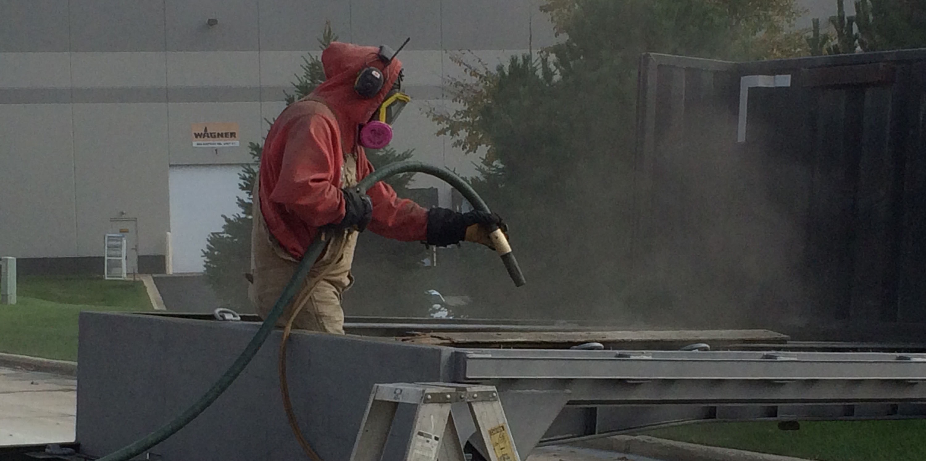 Worker in red jacket operating blasting equipment on industrial components